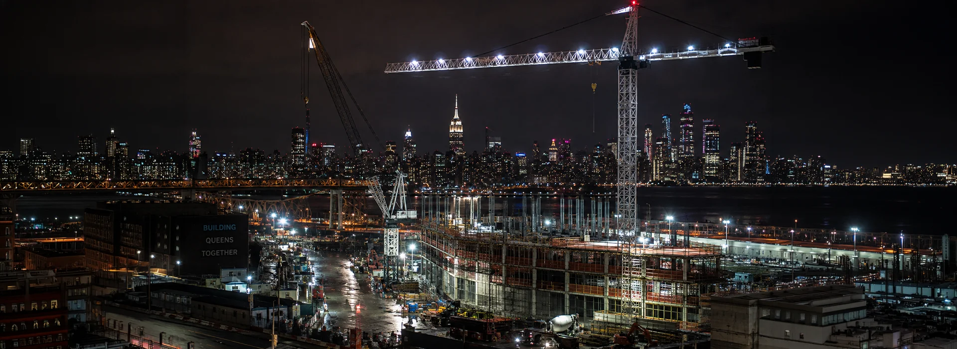 Nighttime construction site in Queens with illuminated cranes and the city skyline behind