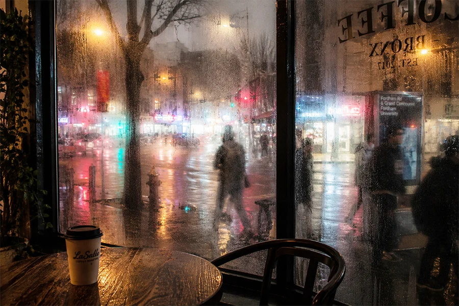 View from inside a Bronx cafe through a rain-streaked window with blurred people passing outside